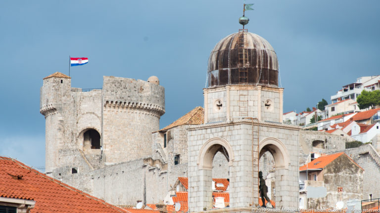 Domed stone bell tower with a crenellated fortress tower and hillside buildings behind it.