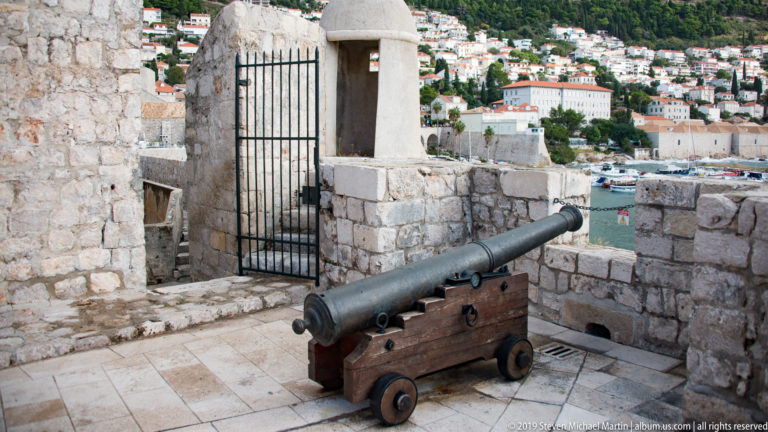 Historic cannon on a wooden carriage beside a stone wall overlooking Dubrovnik harbor.