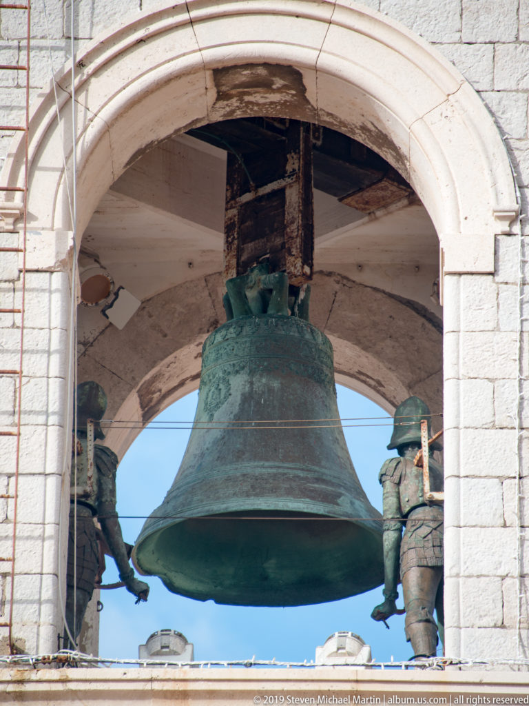 Large green-patinated bronze bell hanging inside a round-arched stone belfry opening.
