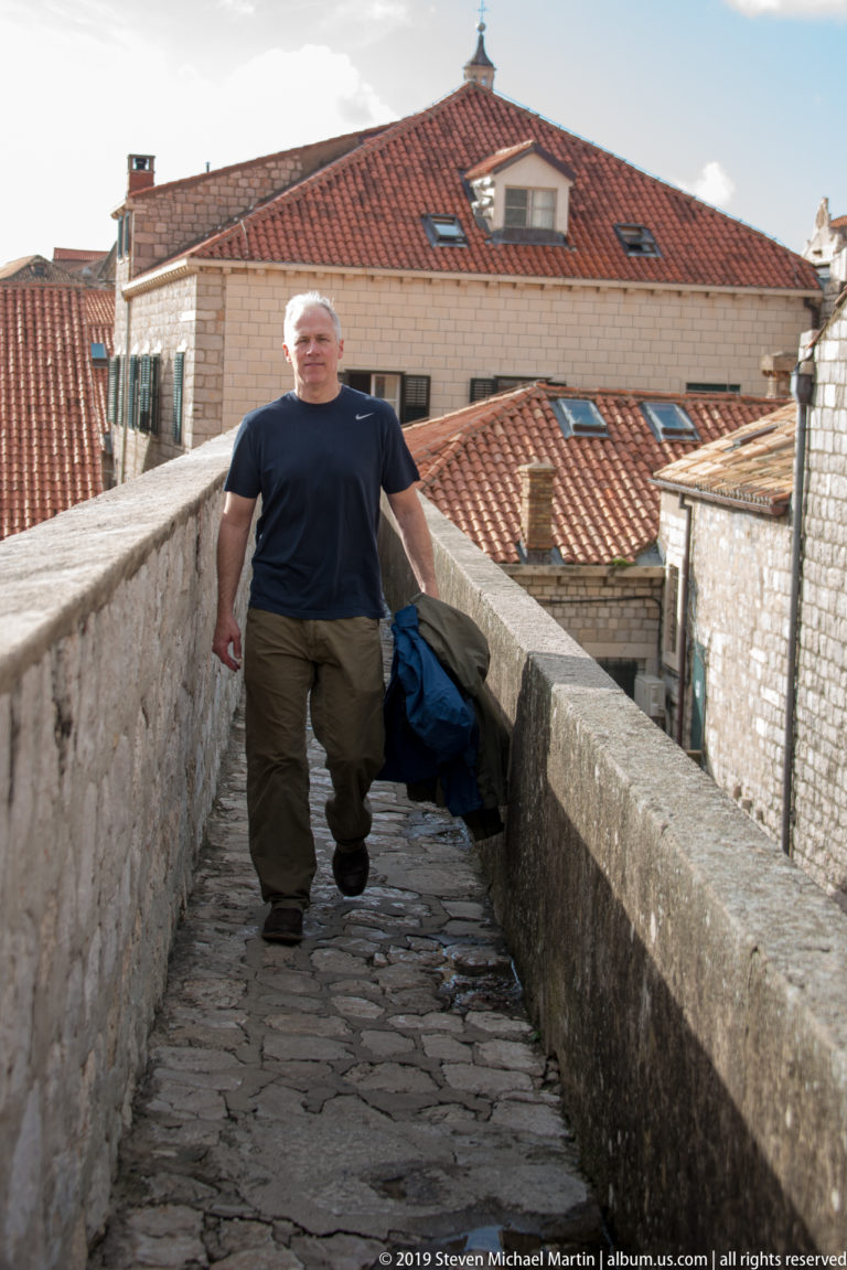 Man standing on a narrow stone wall walk with red-tiled roofs behind him.
