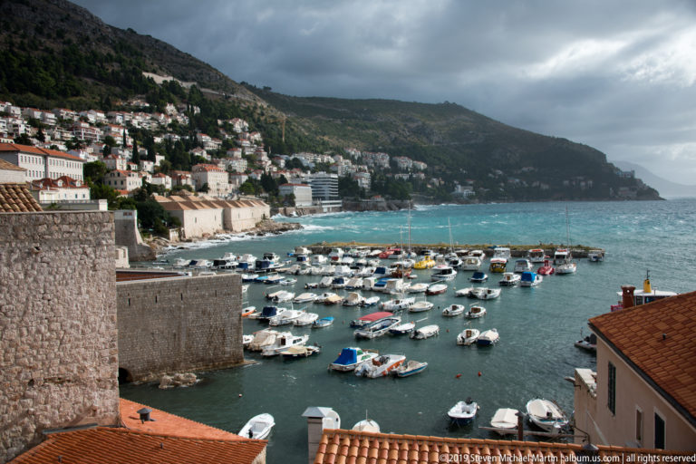 Small boats in Dubrovnik harbor below stone walls and a hillside under dark storm clouds.