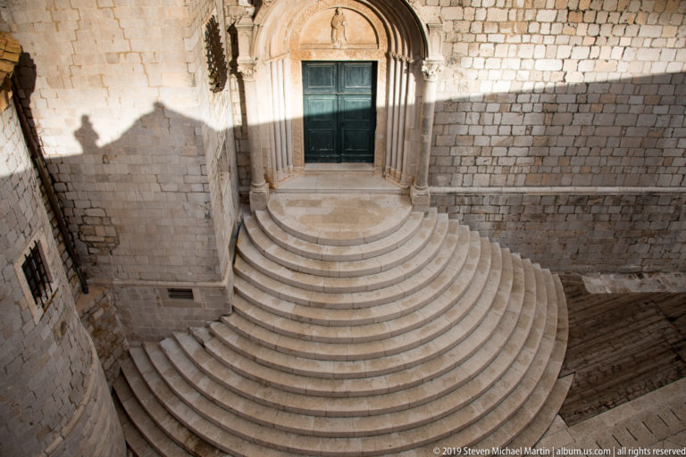 Wide semicircular stone steps leading to an arched doorway in a pale masonry wall.