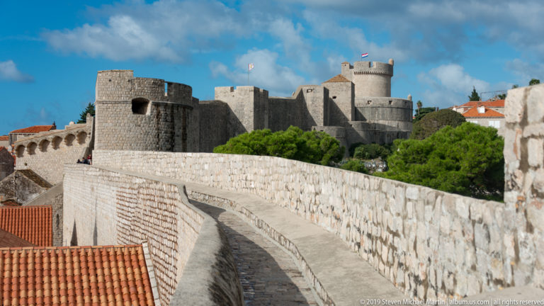 Curving stone wall walk leading toward massive fortified towers under a blue sky.