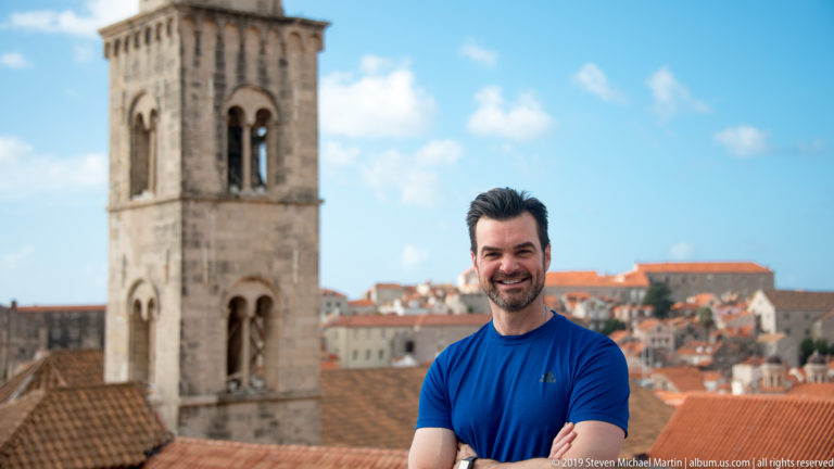 Man in blue shirt standing above red-tiled Dubrovnik roofs beside a pale stone bell tower.