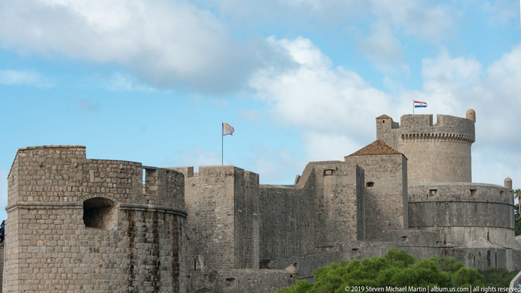 Round Minčeta Tower rises behind overlapping pale stone curtain walls and lower defensive towers beneath a blue sky.