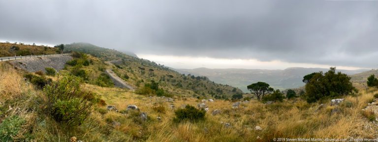 Dry rocky hillside with grasses and shrubs overlooking a hazy Adriatic landscape under low clouds.