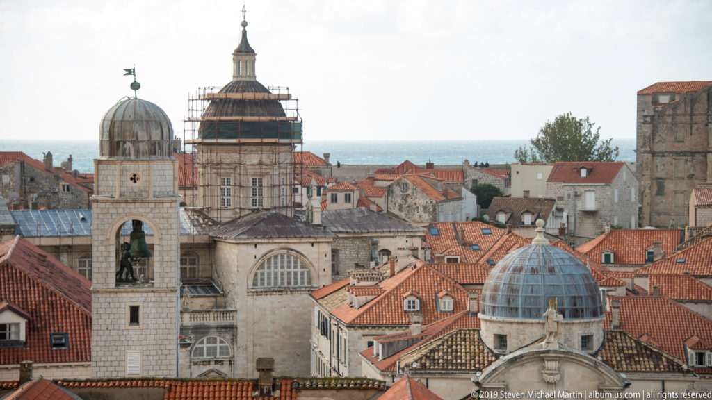 Several church domes and bell towers rise above red Dubrovnik rooftops, including one tower partly covered in scaffolding.