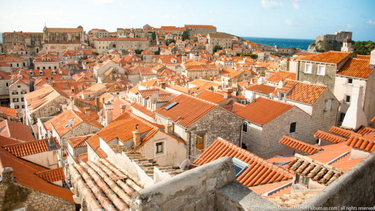 Dense orange-red terracotta roofs and pale stone buildings fill Dubrovnik Old Town beneath a bright blue sky.