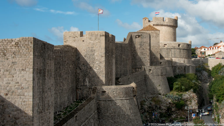 Massive limestone walls and angular defenses lead to the large round Minčeta Tower, with flags flying above the fortifications.