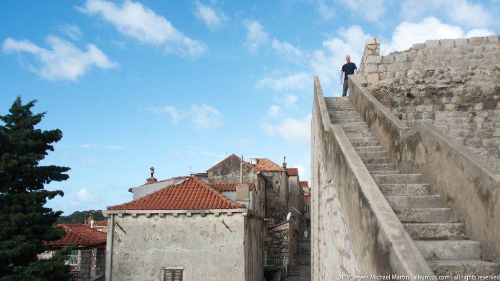 Steep stone staircase climbs beside a weathered city wall toward a visitor at the parapet, with red-tiled roofs below.
