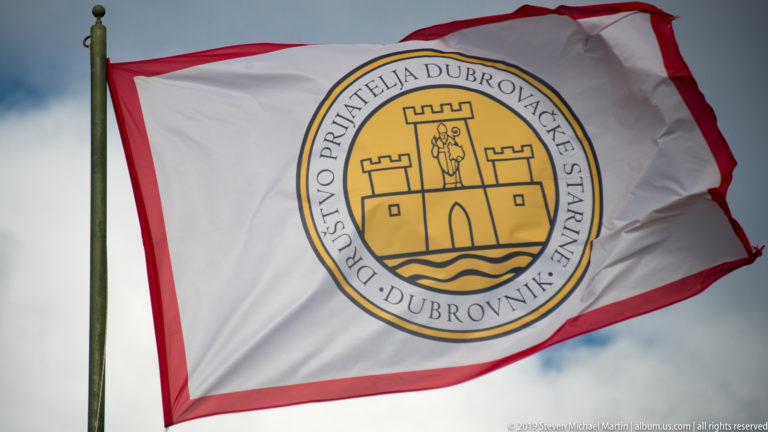 White red-edged flag with a circular yellow emblem, fortified-gate symbol, and Croatian inscription for Dubrovnik’s antiquities society.
