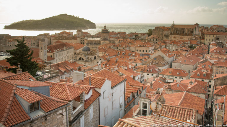 Warm-lit Dubrovnik rooftops surround several church domes and towers, with the Adriatic visible beyond the historic center.