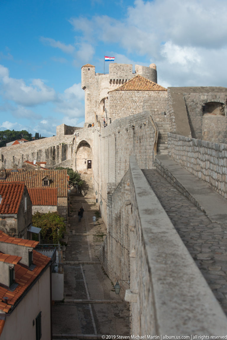 Wide stone rampart leads toward a crenellated tower and wall, with red-tiled roofs closely lining the fortifications.
