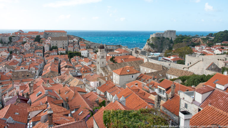 Dubrovnik rooftops fill the foreground while Fort Lovrijenac rises on a rocky cliff beside the blue Adriatic in the distance.
