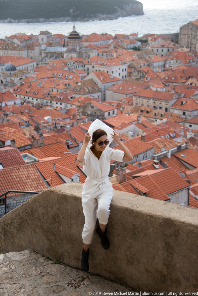 Visitor in white adjusts a hat beside a low stone parapet, with dense red Dubrovnik rooftops spreading behind.