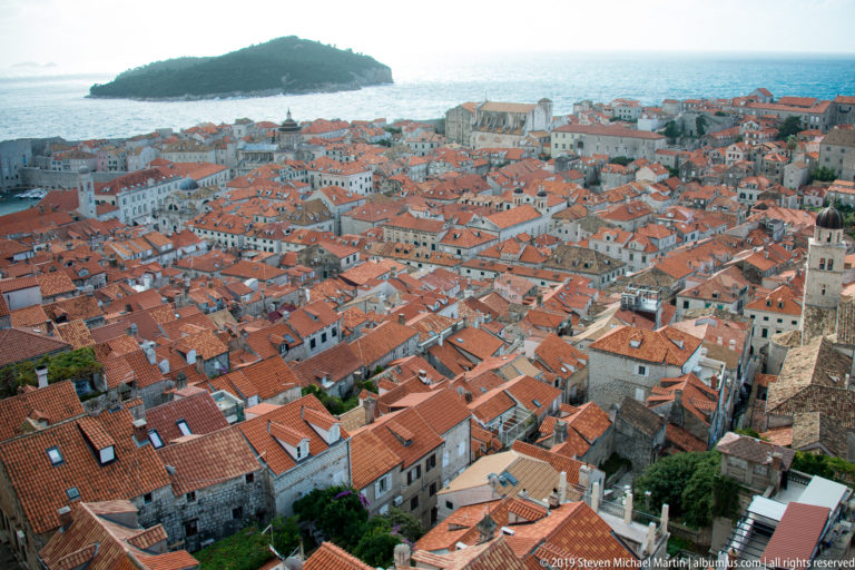 Dense terracotta rooftops and pale stone buildings fill Dubrovnik Old Town, with blue sea and a wooded island beyond.
