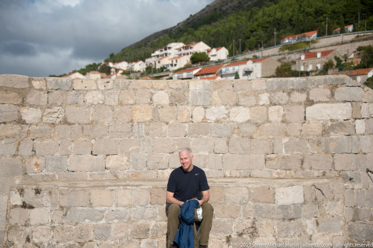 Seated visitor in dark shirt rests against a broad pale stone wall, with hillside houses visible above the parapet.