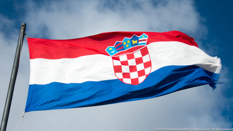 Croatian flag with red, white, and blue bands and checkerboard coat of arms billows against a blue cloudy sky.