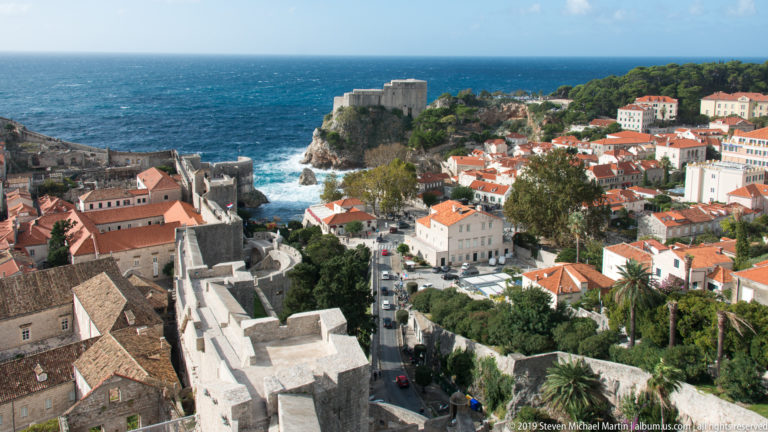 High view from Dubrovnik’s walls over red-roofed buildings and ramparts toward Fort Lovrijenac beside the blue Adriatic.