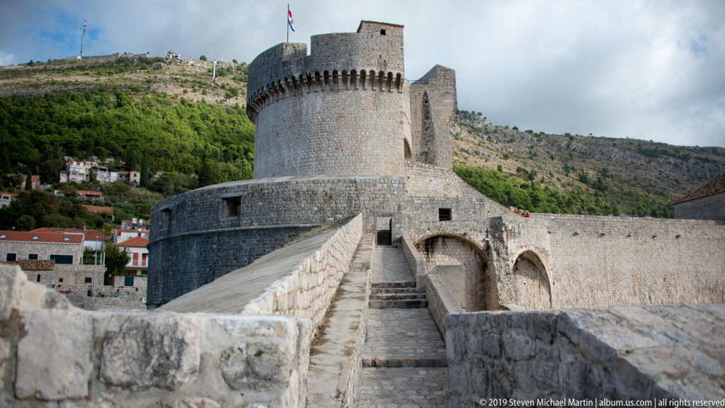 Round Minčeta Tower rises ahead of a narrow stone wall walk, with high masonry defenses and green hillside behind.