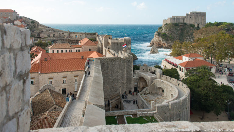 Dubrovnik wall walk curves through towers and red roofs toward Fort Lovrijenac on a rocky cliff above the sea.