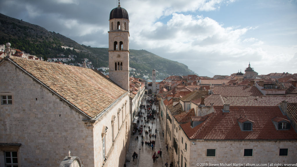 Tall stone bell tower and long tiled roof overlook a crowded pedestrian street and dense red-roofed Dubrovnik buildings.