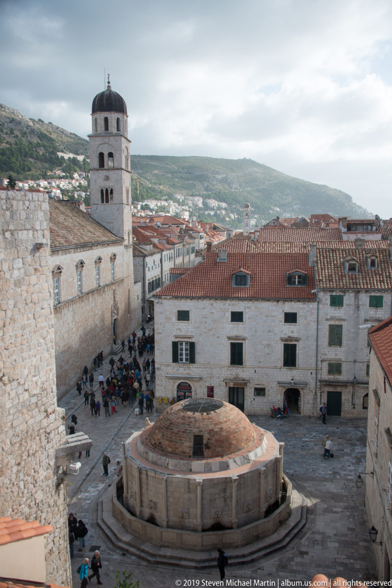 Large polygonal stone fountain with a domed roof stands in a Dubrovnik square, surrounded by pale buildings and pedestrians.