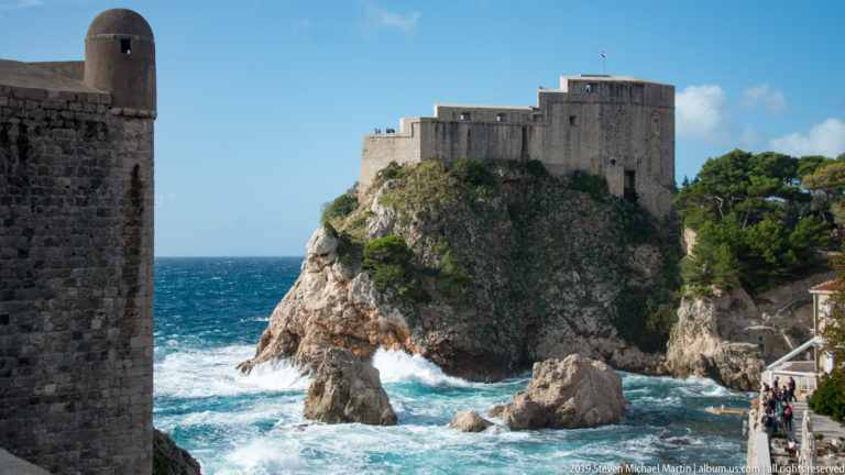 Fort Lovrijenac stands on its rocky promontory, partly framed by a dark city wall with visitors visible at lower right.
