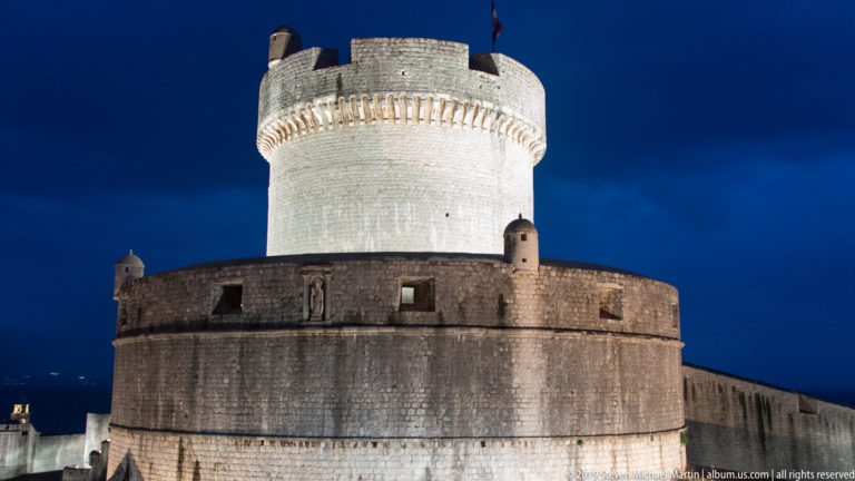 Large floodlit circular defensive tower with corbelled upper stage, small turrets, flag, and adjoining Dubrovnik city wall.