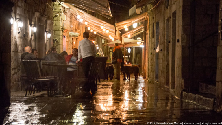 Outdoor diners beneath glowing awnings in a narrow rain-wet stone lane in Dubrovnik’s Old City at night.