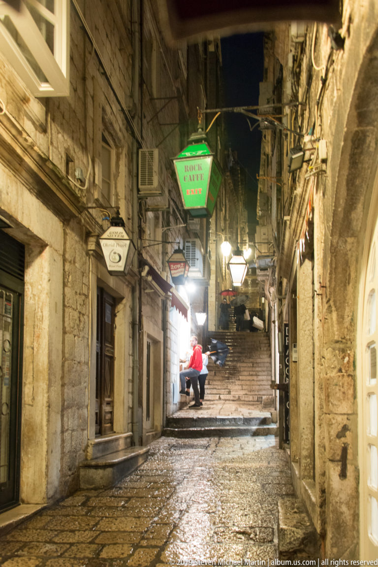 Wet stone lane climbing between old buildings with shop signs, lamps, and steps in Dubrovnik at night.