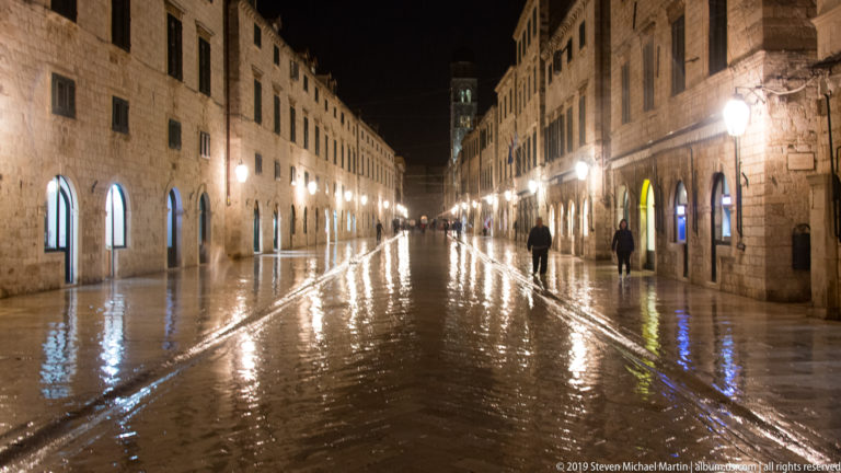 Centered night view along wet Stradun with bright reflections, stone façades, street lamps, and sparse pedestrians.
