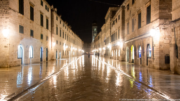 Long rain-wet view down Stradun between historic stone façades, glowing shopfront arches, lamps, and a few pedestrians.