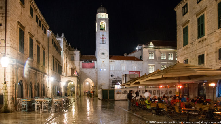Rain-wet street with café diners and empty tables leading toward Dubrovnik’s illuminated City Bell Tower.