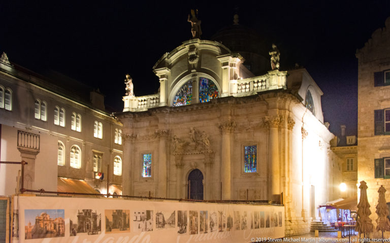 Illuminated Church of St. Blaise with Baroque façade, rooftop statues, stained glass, and exhibition panels in front.