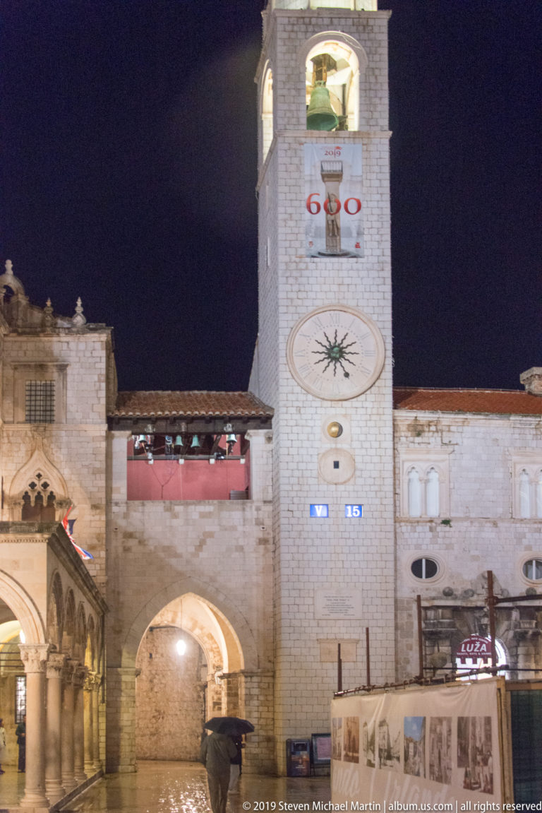 Dubrovnik City Bell Tower with illuminated belfry, oval clock, Orlando banner, and archway at Luža Square.