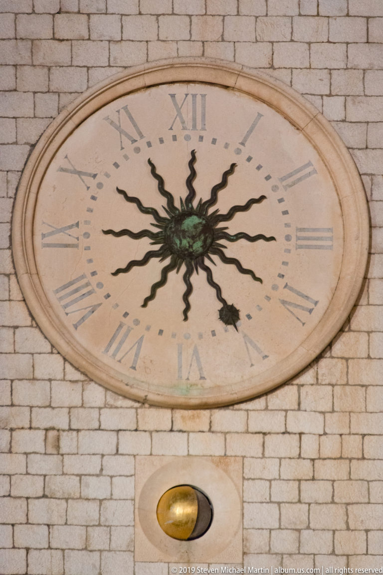 Oval historic clock with Roman numerals and dark radiating hands mounted on pale stone masonry.