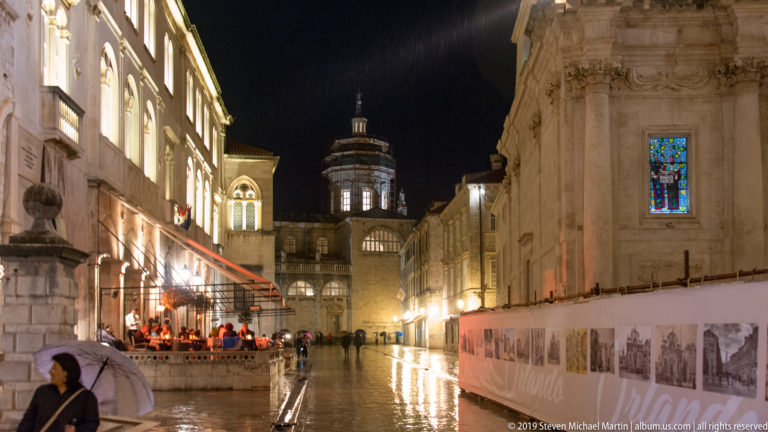 Rainy Dubrovnik street with café diners, pedestrians, temporary exhibition panels, and illuminated cathedral rising ahead.