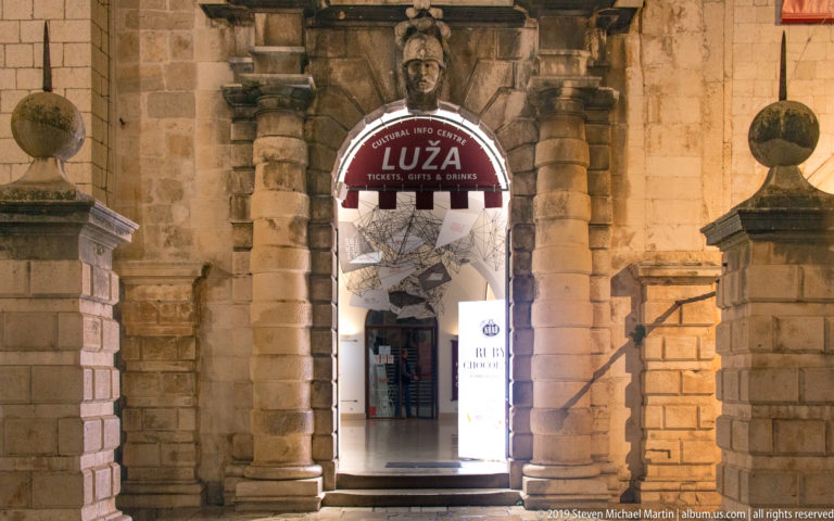 Illuminated arched doorway marked LUŽA, set in rusticated stone with visitor-services signs and sculptural decoration.