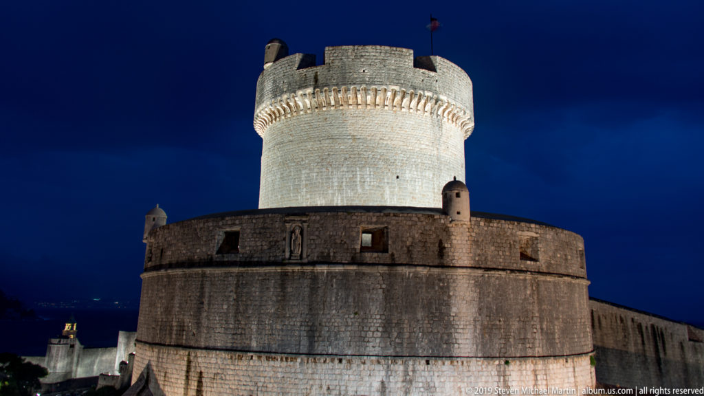 Floodlit circular fortification with tall cylindrical upper tower, corbelled parapet, sentry turrets, and adjoining city wall.