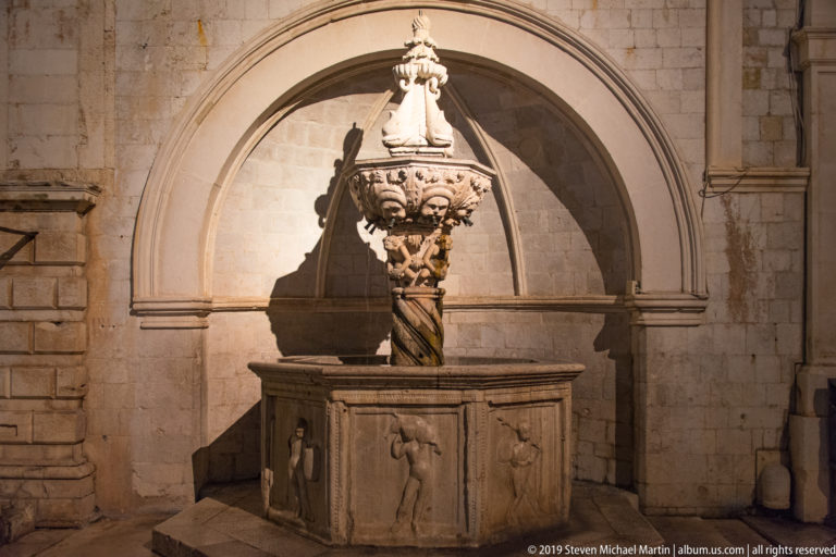 Small Onofrio Fountain beneath a stone arch, with carved central column, basin, sculpted faces, and ornamental finial.