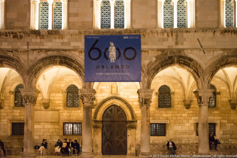 Rector’s Palace arcade with carved columns, pointed doorway, seated visitors, and Orlando anniversary banner at night.
