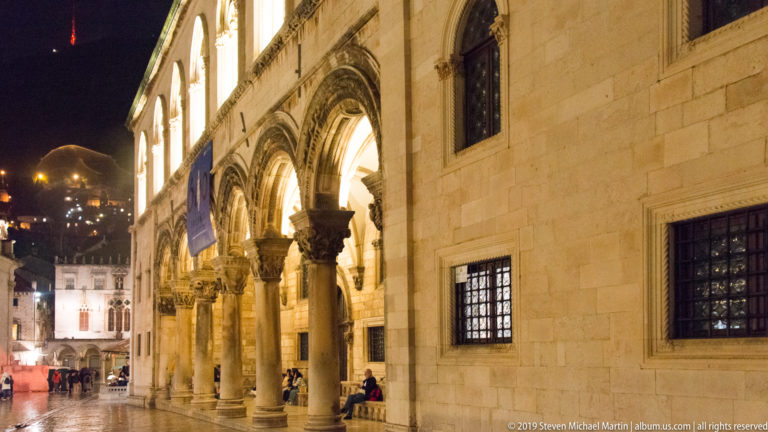 Rector’s Palace arcade with carved columns and arches beside a wet square, with civic buildings receding beyond.