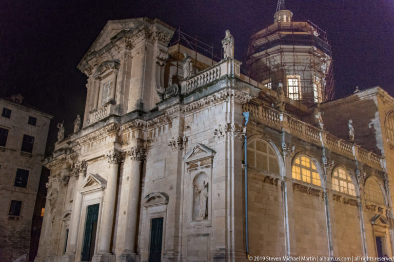 Oblique illuminated view of Dubrovnik Cathedral showing façade, side wall, rooftop statues, windows, and scaffolded dome.