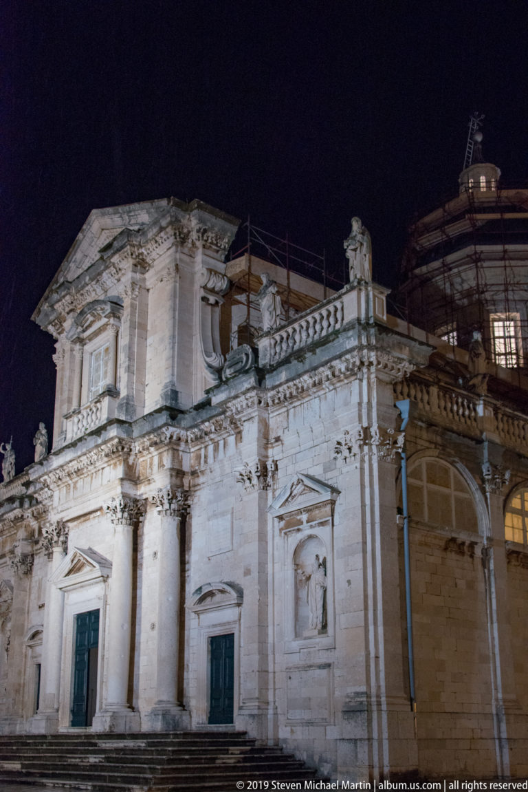 Illuminated Baroque façade of Dubrovnik Cathedral with statues, pilasters, balustrade, and scaffolded dome behind.