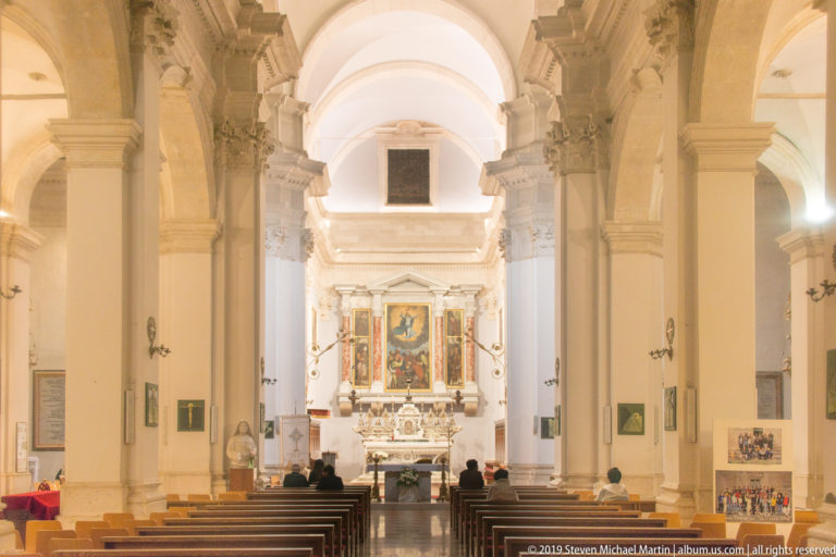 Long church nave with tall piers, arches, rows of pews, vaulted ceiling, visitors, and illuminated marble altar.