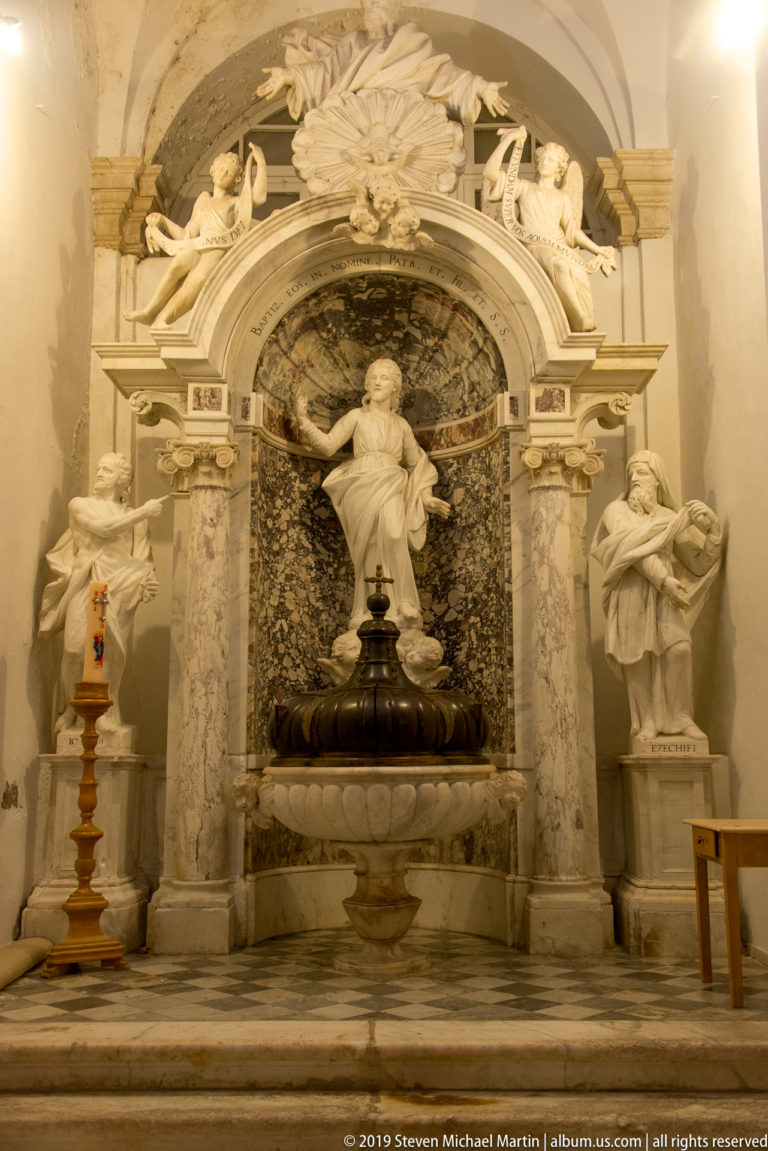 Marble niche with central robed figure above a dark basin, flanked by statues and ornate architectural framing.