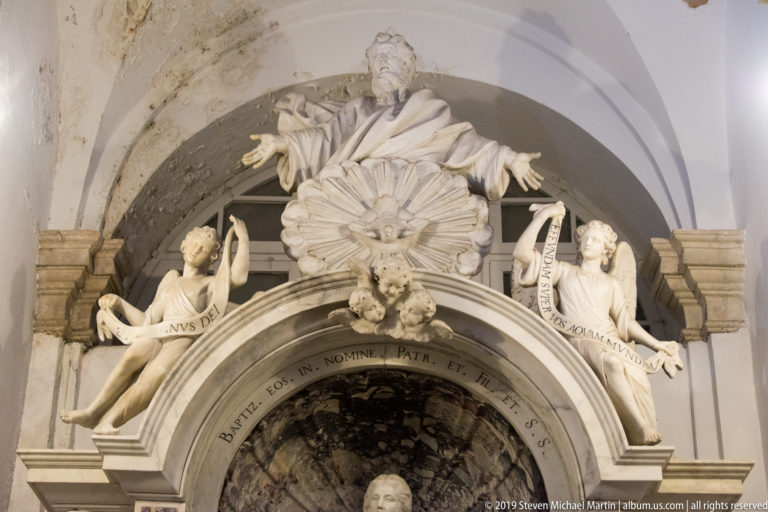 Baroque marble monument in an arched recess with central robed figure, flanking figures, inscribed ribbons, and sculpted clouds.