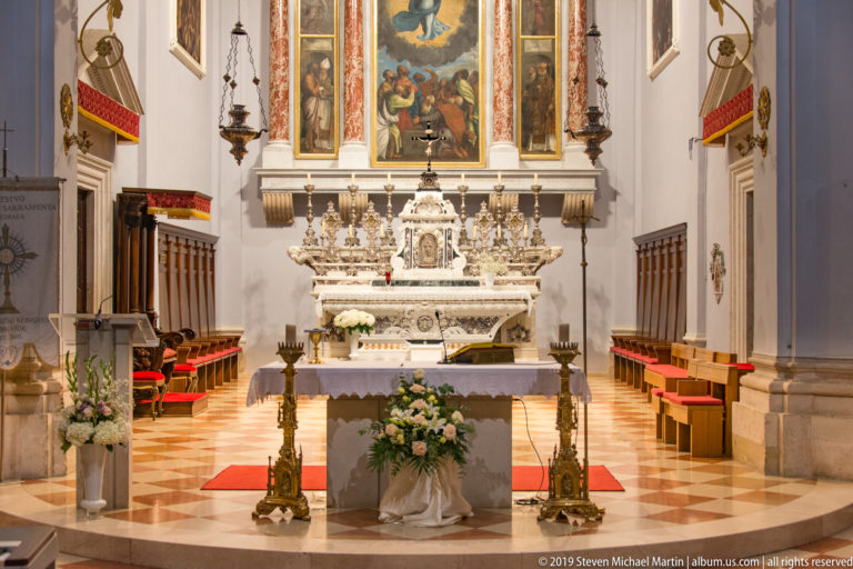 Marble high altar with tabernacle, central religious painting, side panels, candlesticks, flowers, and choir stalls.