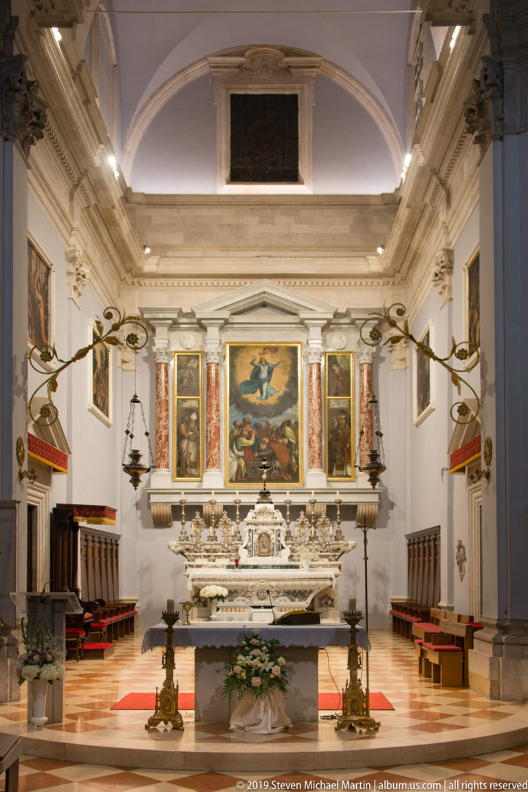 Bright church sanctuary with marble altar, large central painting, flanking panels, hanging lamps, choir stalls, and flowers.